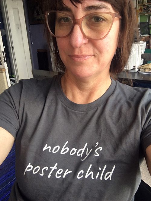 A confident-looking young woman takes a selfie while wearing a "nobody's poster child" tee shirt.